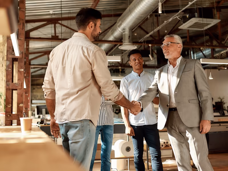 Two cheerful men shaking hands and smiling while standing with coworkers in the modern office