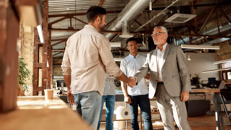 Two cheerful men shaking hands and smiling while standing with coworkers in the modern office