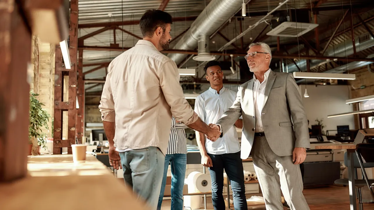 Two cheerful men shaking hands and smiling while standing with coworkers in the modern office